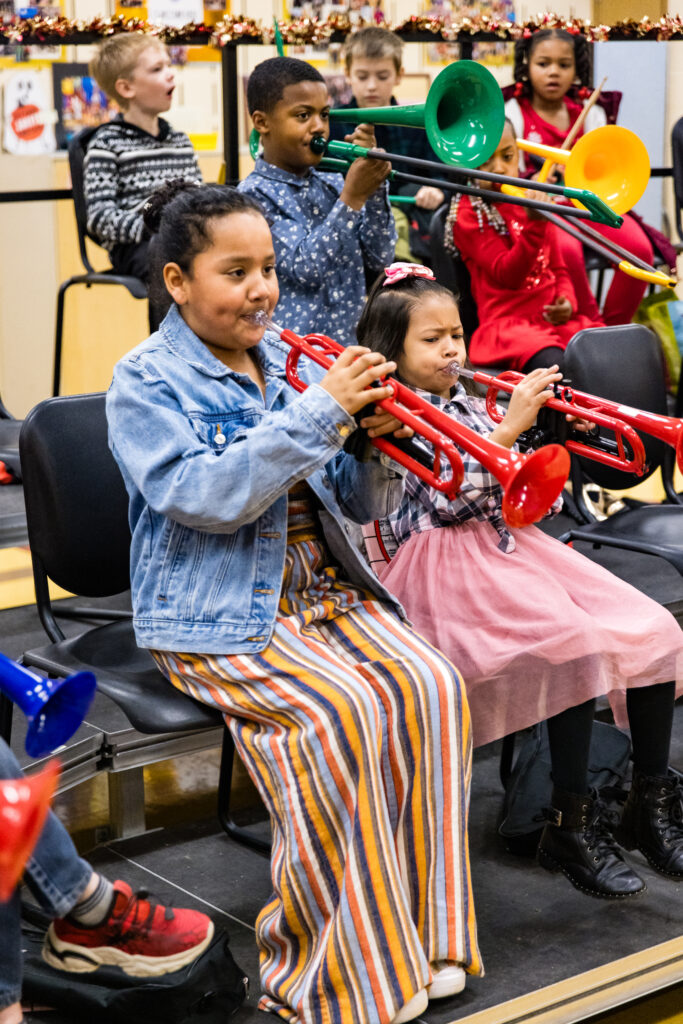 Young students practicing colorful plastic trumpets on risers in a classroom.