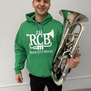 Man wearing green hoodie with white RCB logo, holding a euphonium and smiling